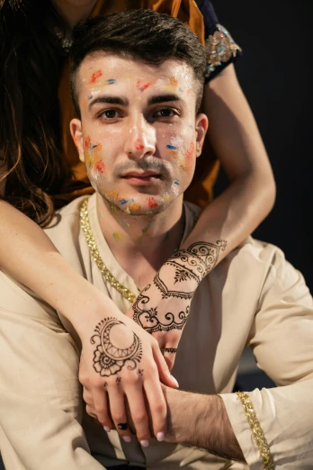 a man and a woman sitting next to each other, a tattoo, trending on pexels, renaissance, tribal facepaint, hamsa hand, beautiful young man, glitter