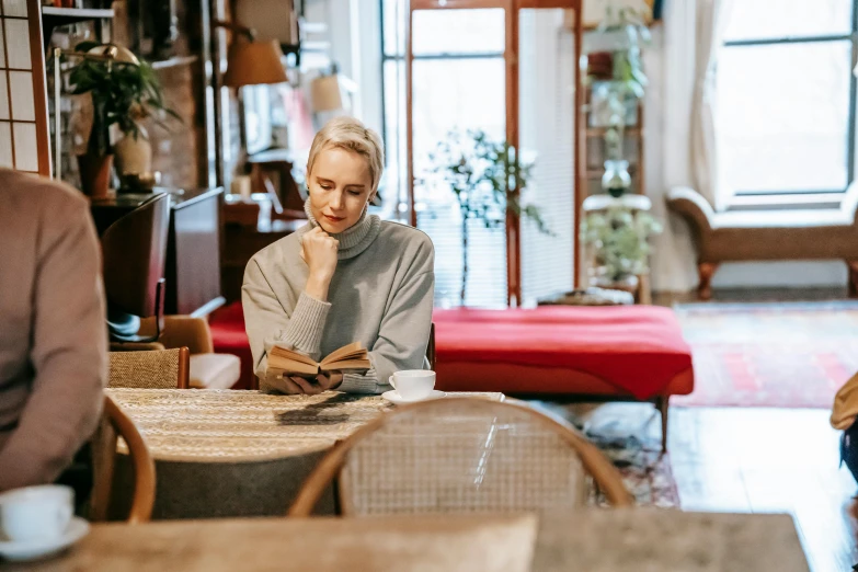 a woman sitting at a table reading a book, by Emma Andijewska, pexels contest winner, sitting in a lounge, looking smart, a blond, bookshops