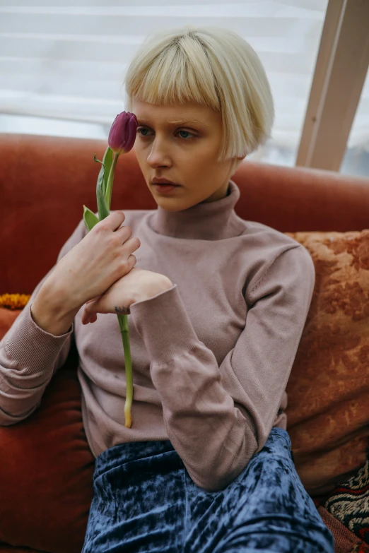 a woman sitting on a couch holding a flower, inspired by Elsa Bleda, trending on pexels, aestheticism, wearing turtleneck, pale hair, russian girlfriend, promotional image