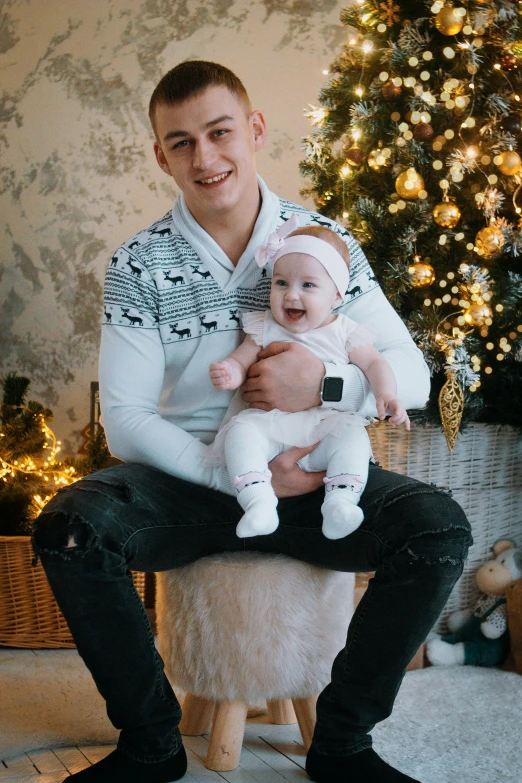 a man holding a baby in front of a christmas tree, a picture, dasha taran, dressed casually, portrait image, relaxing