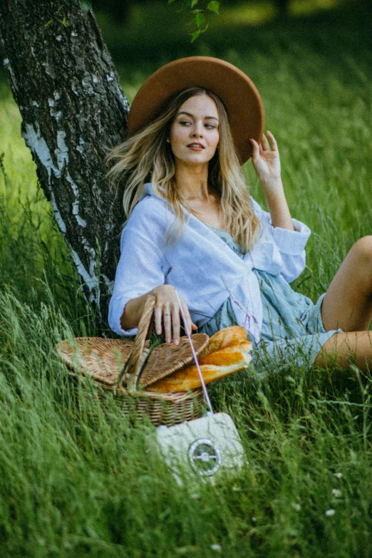 a woman sitting in the grass next to a tree, carrying a saddle bag, holding a baguette, photoshoot for skincare brand, food