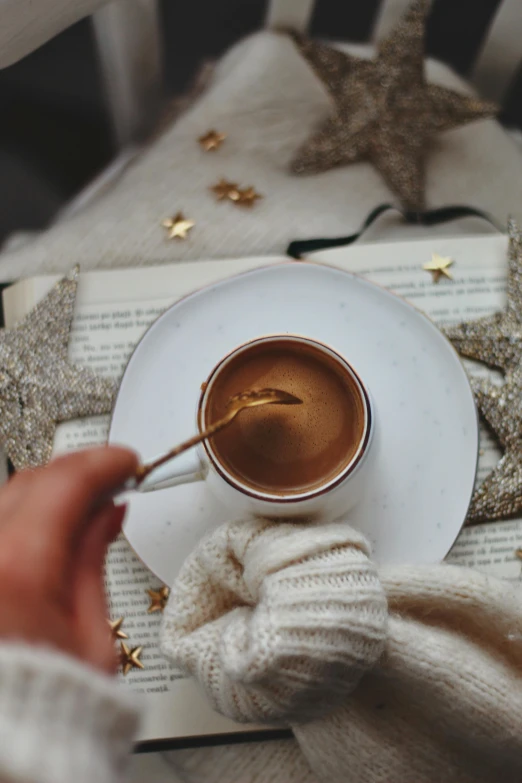 a person holding a spoon over a cup of coffee, by Julia Pishtar, pexels contest winner, romanticism, tiny stars, brown sweater, thumbnail, holiday