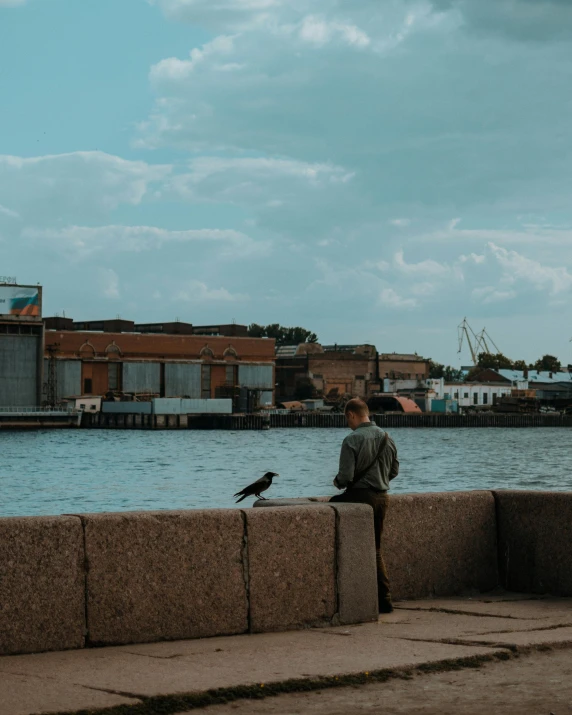 a person sitting on a wall next to a body of water, by Jan Tengnagel, pexels contest winner, city docks, bird on his shoulder, low quality photo, calmly conversing 8k