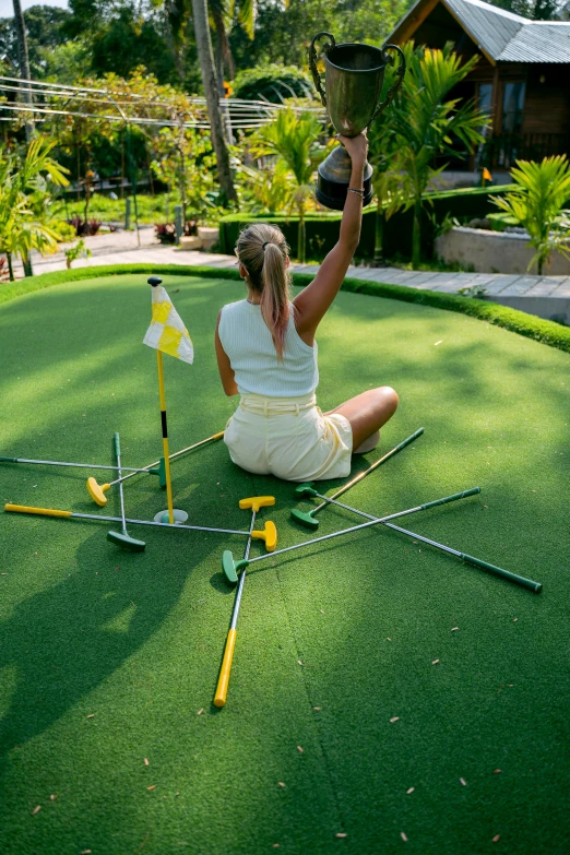 a woman sitting on top of a putting green, set pieces, dabbing, yellow carpeted, lush surroundings