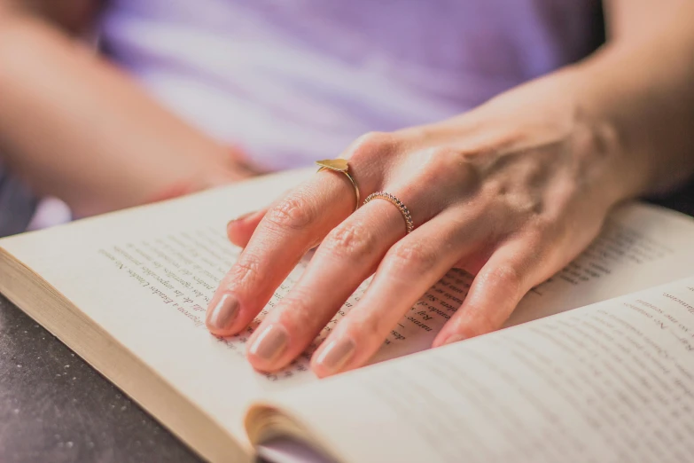a close up of a person reading a book, wearing two metallic rings, wearing gold jewellery, raised hand, gold and purple
