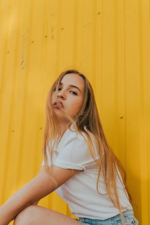 a woman sitting on the ground in front of a yellow wall, inspired by Elsa Bleda, trending on pexels, realism, long golden hair, dressed in a white t shirt, 15081959 21121991 01012000 4k, headshot profile picture