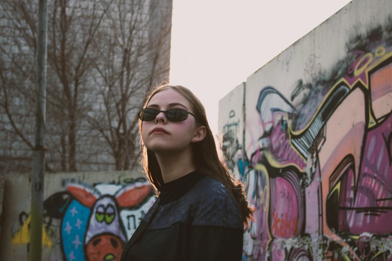 a woman standing in front of a graffiti covered wall, an album cover, inspired by Elsa Bleda, pexels contest winner, sunglasses, portrait sophie mudd, looking up, low quality photo