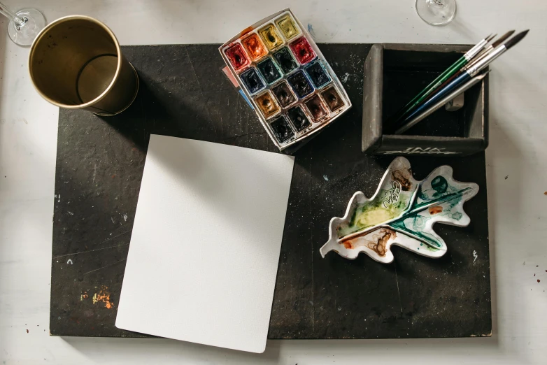 a piece of paper sitting on top of a table next to a cup of coffee, a watercolor painting, inspired by Kyffin Williams, pexels contest winner, paintbrush and palettes, matte white paint, painting on black canvas, children's artwork