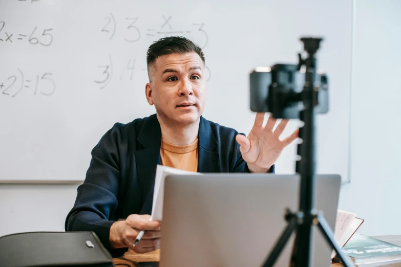 a man sitting at a table in front of a laptop, unsplash, academic art, sitting in front of a microphone, maths, camera footage, teacher