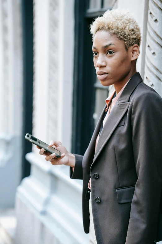 a woman standing in front of a building holding a cell phone, trending on pexels, wearing a worn out brown suit, black young woman, androgynous person, 1 4 5 0