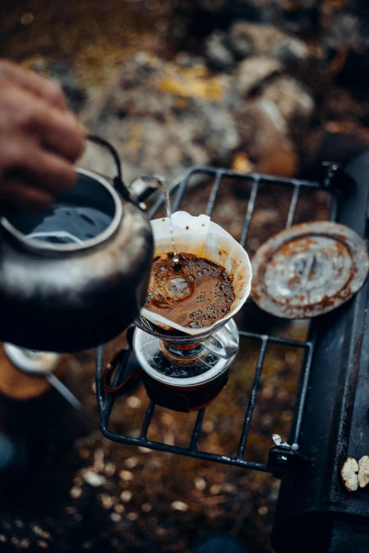 a person holding a kettle over a cup of coffee, by Jan Tengnagel, trending on unsplash, outdoor campfire pit, “ iron bark, coffee stain, patterned