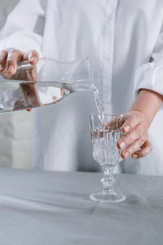 a person pouring water into a glass on a table, by Isabel Naftel, trending on unsplash, renaissance, made of crystal, linen, detailed product image, grey