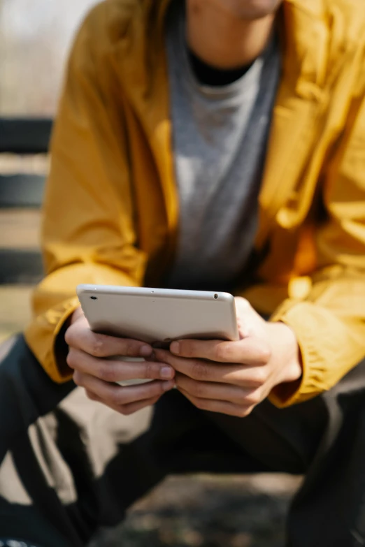 a man sitting on a bench using a cell phone, wearing a yellow hoodie, ipad pro, zoomed in, holding controller