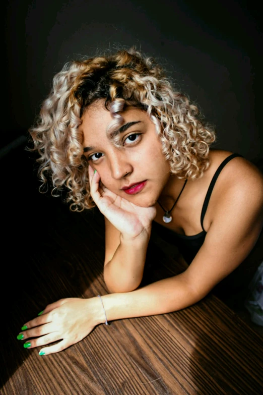 a woman sitting at a table with her hand on her chin, an album cover, inspired by Elsa Bleda, pexels contest winner, photorealism, silver curly hair, standing with a black background, low quality photo, 🤤 girl portrait