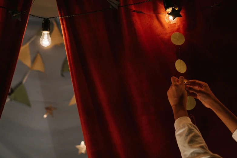 a person reaching up to a string of lights, by Julia Pishtar, pexels contest winner, red curtains, children playing with pogs, gold decorations, holding a lantern