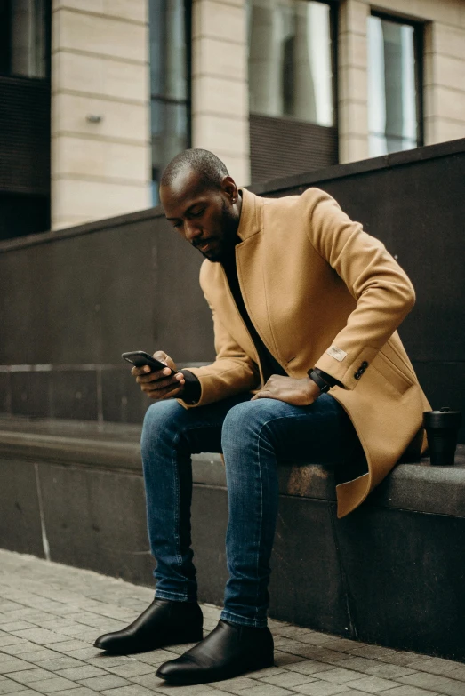 a man sitting on a ledge looking at his cell phone, trending on pexels, happening, light brown coat, man is with black skin, impeccably dressed, black and orange coat