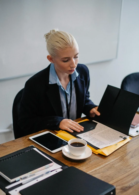 a woman sitting at a table with a laptop and a cup of coffee, wearing causal black suits, school curriculum expert, multiple stories, holding a clipboard