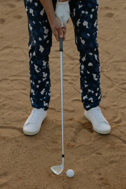 a man standing on top of a sandy beach holding a golf club, by Jan Tengnagel, unsplash, op art, large pants, desert camouflage, close up image