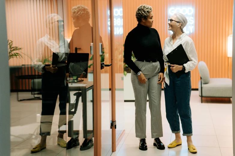 a couple of women standing next to each other, by Carey Morris, pexels contest winner, in the office, grey pants and black dress shoes, peach and goma style, non-binary