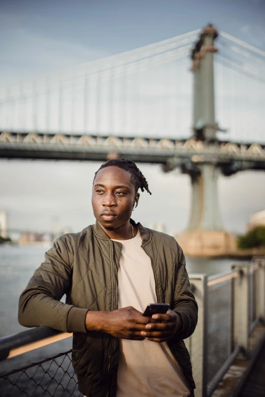 a man standing in front of a bridge looking at his cell phone, a picture, by Joseph Severn, with brown skin, brooklyn background, he is about 20 years old | short, postprocessed