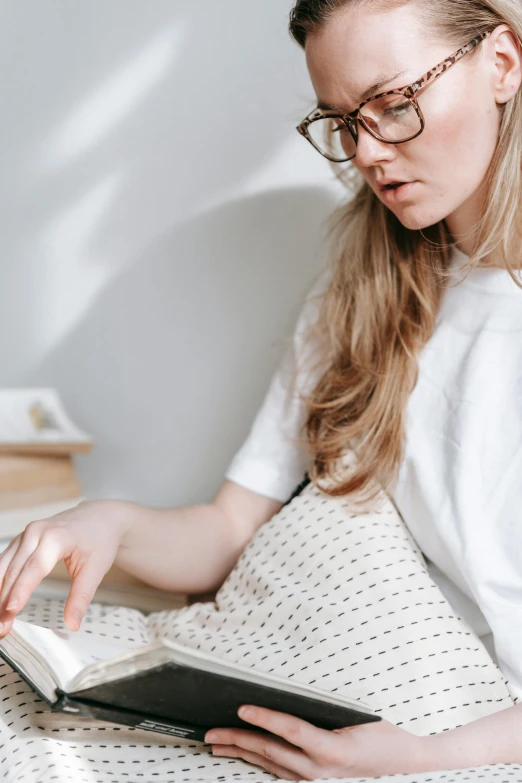 a woman sitting on a bed reading a book, trending on pexels, happening, white shirt and grey skirt, maternity feeling, with glasses on, freckled pale skin