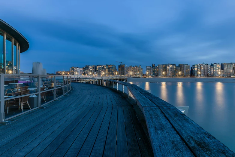 a dock next to a body of water with buildings in the background, by Matthias Stom, pexels contest winner, blue hour, boardwalk, panorama, at the seaside