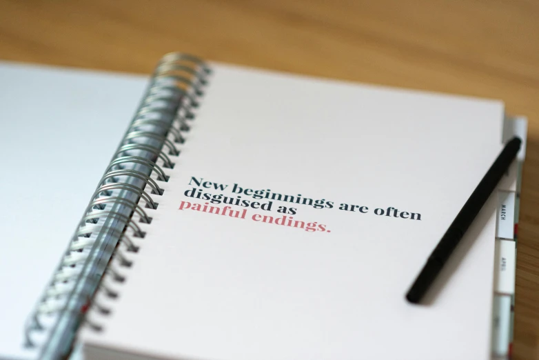 a notebook with a pen sitting on top of it, by Gen Paul, new objectivity, brand colours are red and blue, inspirational quote, endings, on a pale background