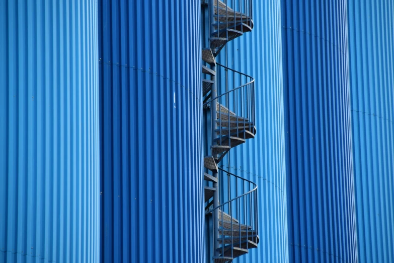 a spiral staircase going up the side of a blue building, industrial plant environment, telephoto shot, shipping containers, abstract conceptual