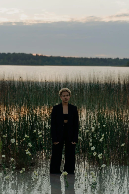 a woman standing in the middle of a body of water, an album cover, by Attila Meszlenyi, unsplash, hurufiyya, in a black suit, standing in tall grass, sitting in front of a lake, dusk