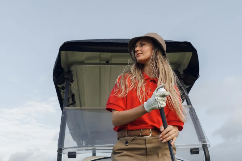 a woman standing in front of a golf cart, pexels contest winner, plain uniform sky at the back, red dress and hat, brown, work clothes