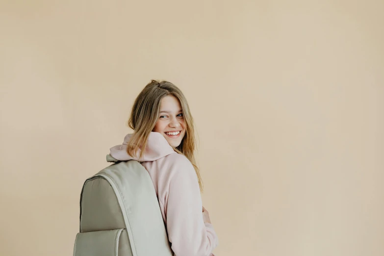 a woman with a backpack standing in front of a beige wall, by Carey Morris, pexels contest winner, silver，ivory, happy girl, silicone skin, background image