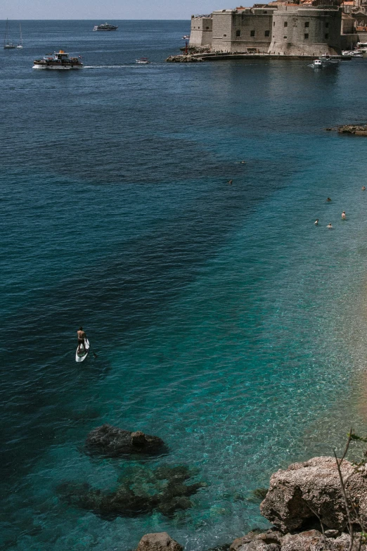 a group of people standing on top of a beach next to a body of water, piroca, deep blue water, standing on surfboards, dubrovnik