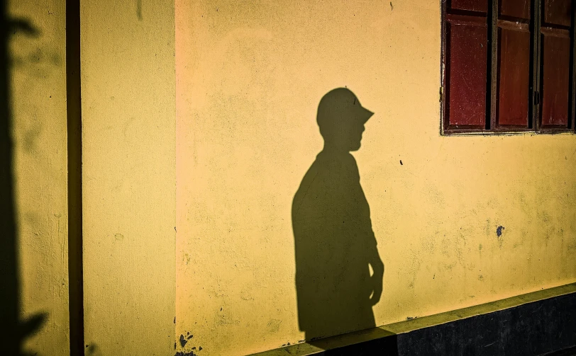 a shadow of a man standing in front of a building, by Eglon van der Neer, pexels contest winner, yellow cap, childhood friend, yellow backdrop, 4 0 years old man