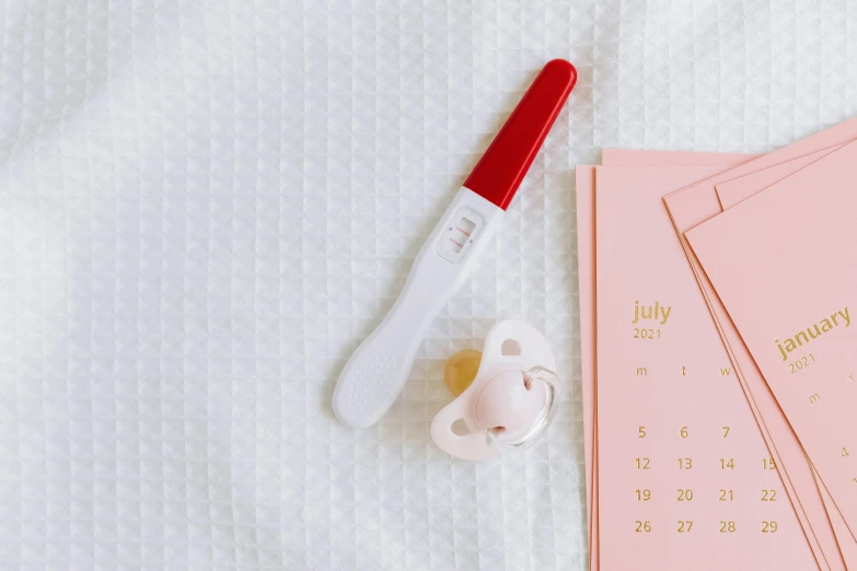 a baby pacifier sitting on top of a bed next to a calendar, trending on pexels, red scales, minimal pink palette, spoon placed, pregnant