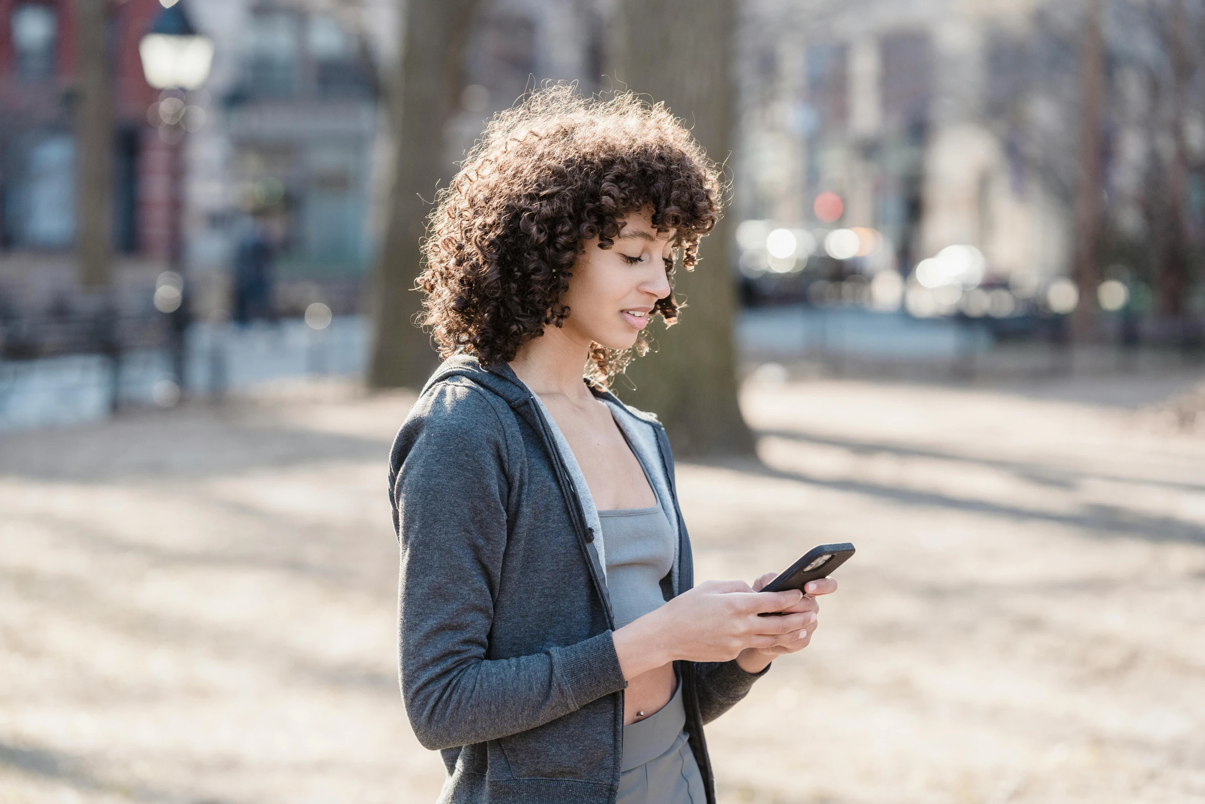 a woman standing in a park looking at her cell phone, trending on pexels, happening, brown curly hair, avatar image, people walking around, high resolution image