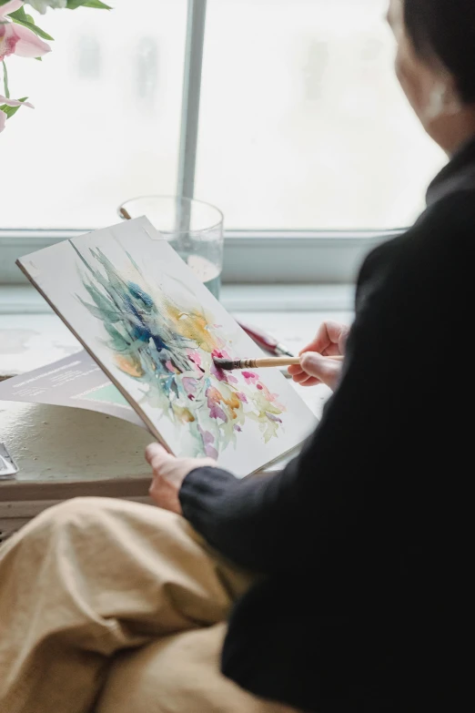 a woman sitting in a chair reading a book, a watercolor painting, inspired by artist, trending on unsplash, process art, on a white table, holding a paintbrush in his hand, botanicals, stands at a his easel