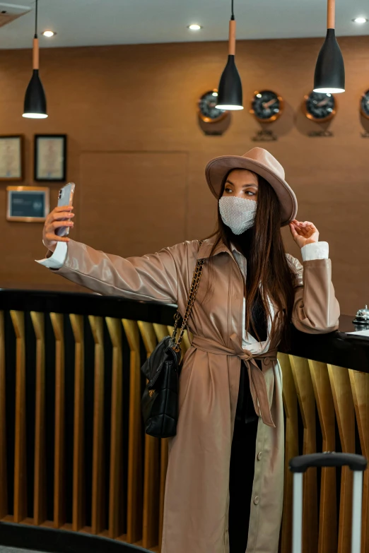 a woman wearing a face mask taking a selfie, trending on pexels, happening, light brown trenchcoat, in a hotel hallway, brown hat, thumbnail