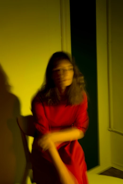 a woman in a red dress leaning against a wall, a picture, by Nathalie Rattner, night vision very blurry, with yellow cloths, her face is in shadow, red room