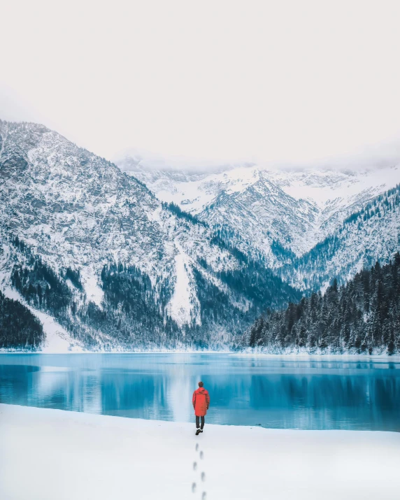 a person walking across a snow covered field next to a lake, pexels contest winner, beach is between the two valleys, red sweater and gray pants, lake blue, centered in portrait