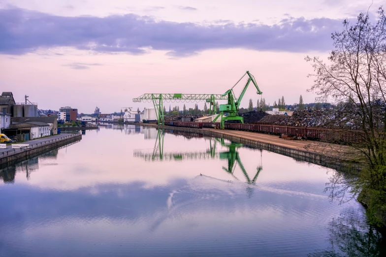 a train traveling down a train track next to a body of water, a photo, inspired by August Lemmer, pexels contest winner, huge machine cranes, in a gentle green dawn light, city docks, water reflection