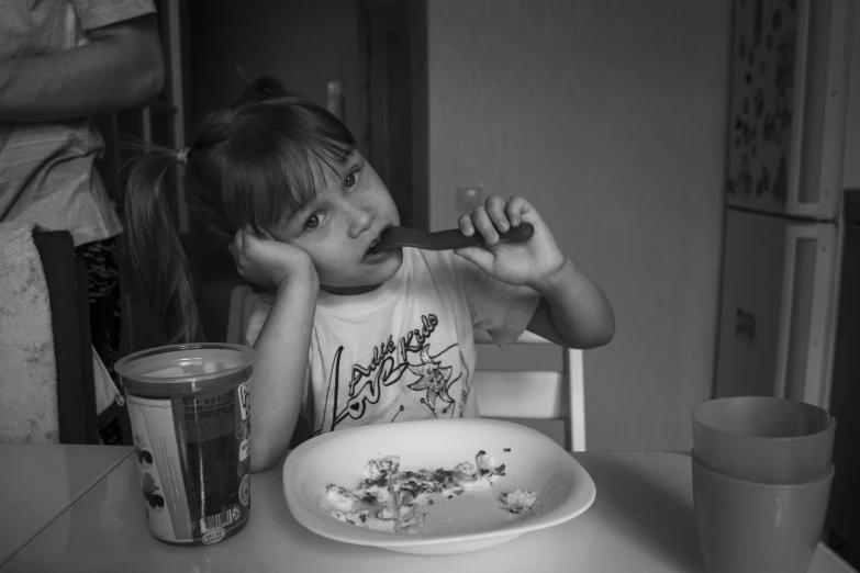 a little girl sitting at a table with a plate of food, a black and white photo, alexey egorov, eating ice cream, at home, impasto