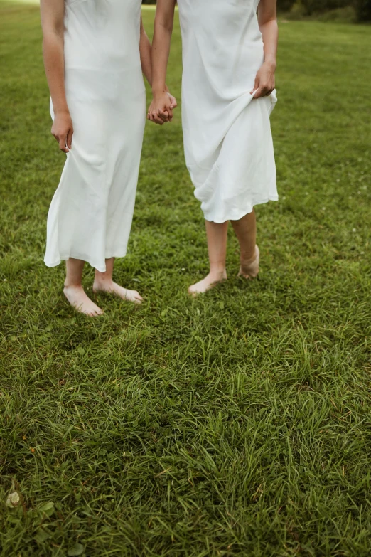 a couple of women standing on top of a lush green field, exposed toes, wearing white cloths, non-binary, uncropped