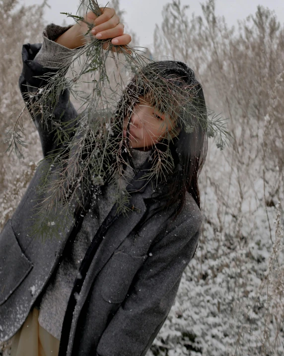 a woman holding a bunch of branches in the snow, unsplash, process art, grey turtleneck coat, androgynous male, gemma chen, greens)