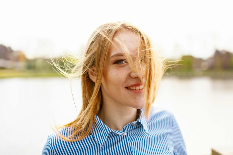 a woman standing in front of a body of water, winking at the camera, golden hair blowing the wind, wearing stripe shirt, avatar image