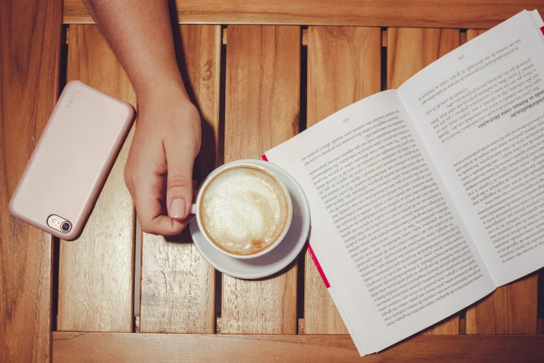 a person holding a cup of coffee next to an open book, happening, 9 9 designs, a wooden, fiction, on a table
