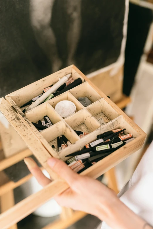 a close up of a person holding a box with pens, a charcoal drawing, by karolis strautniekas, trending on unsplash, wooden cabinet, makeup, wooden crates, white wall coloured workshop