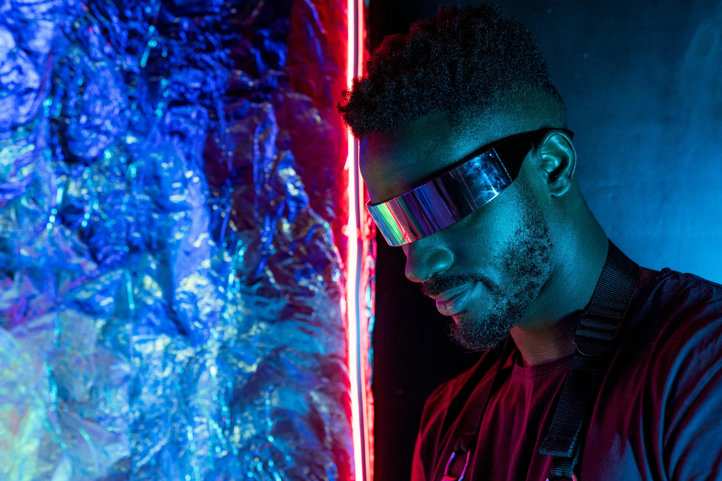 a man standing in front of a wall with neon lights, afrofuturism, futuristic glasses lenses, red reflective lens, holographic blade!, mirror shades