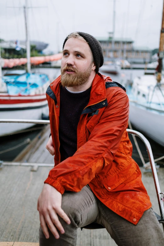 a man with a beard sitting on a boat, a portrait, inspired by Jóhannes Sveinsson Kjarval, unsplash, orange jacket, wearing a red backwards cap, wearing jacket, harbour