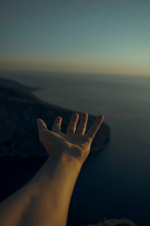 a person standing on top of a cliff next to the ocean, by Alexis Grimou, symbolism, close-up of thin soft hand, lofi, flying shot, during dawn