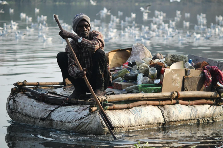 a man sitting on a raft in the middle of a body of water, flickr, environmental art, scattered rubbish and debris, india, an old man with 7 yellow birds, slide show